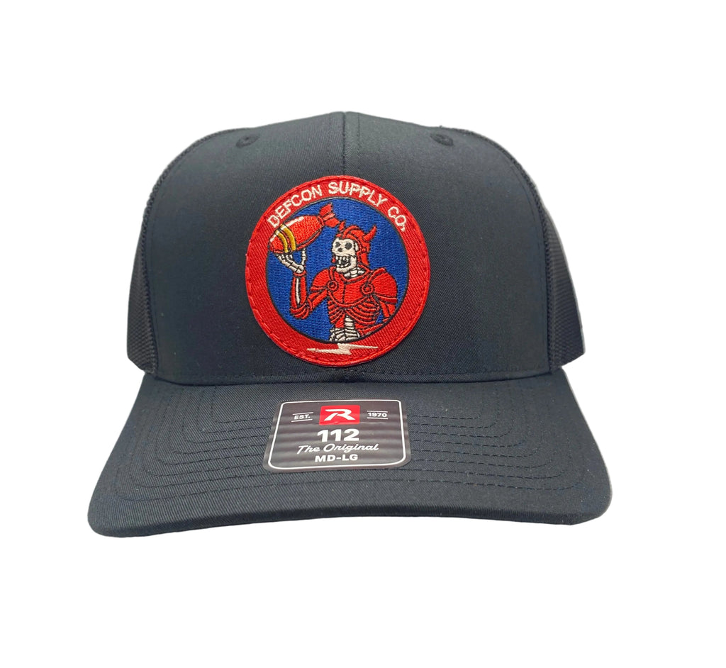 A dark black trucker hat with a curved brim, mesh back, and a circular embroidered front patch featuring a skeleton wearing red armor, holding a bomb, and accented with red, white, and blue design.