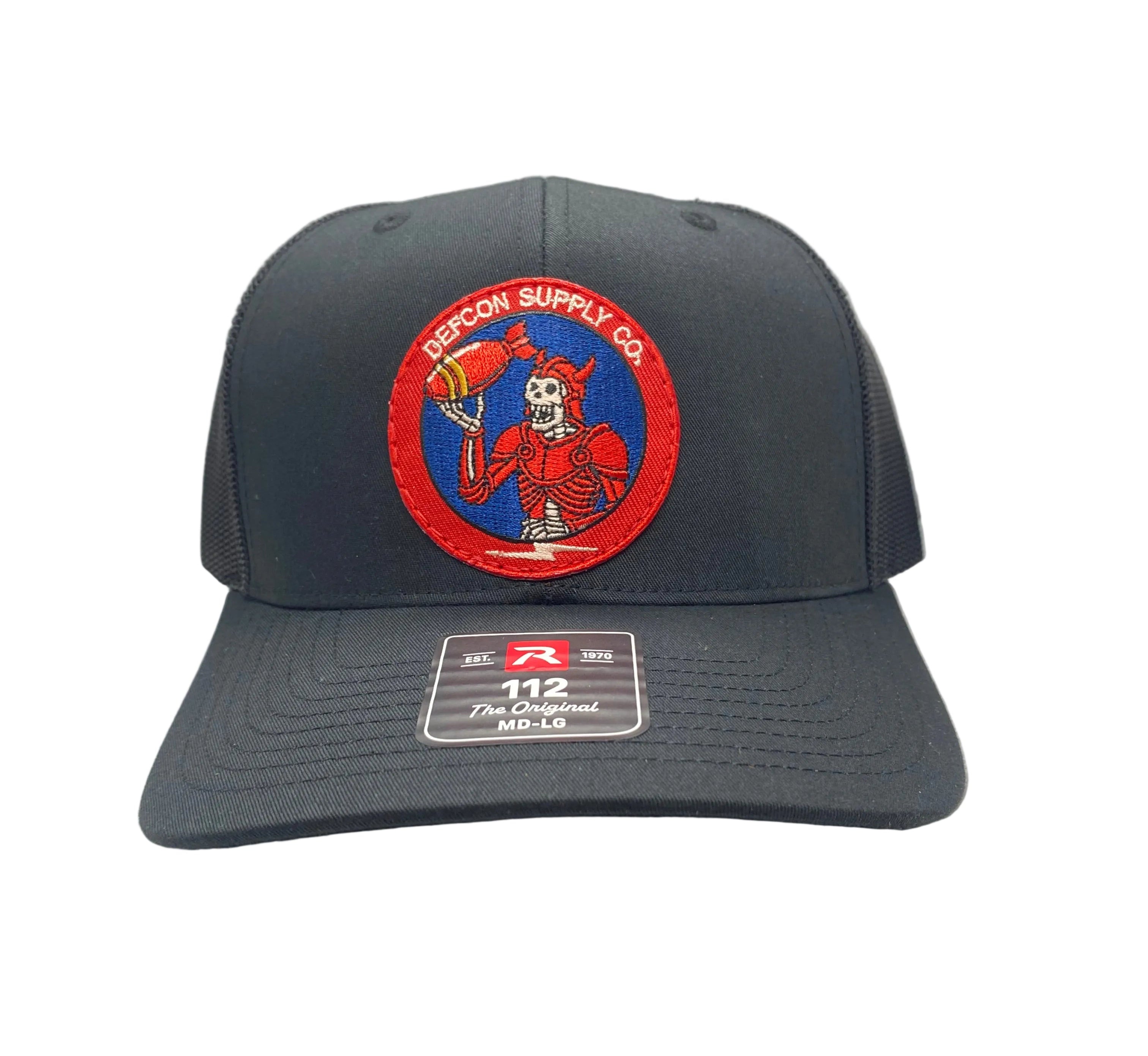 A dark black trucker hat with a curved brim, mesh back, and a circular embroidered front patch featuring a skeleton wearing red armor, holding a bomb, and accented with red, white, and blue design.