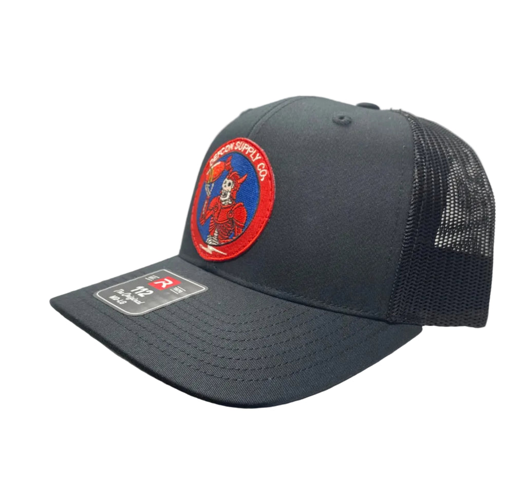 A dark black trucker hat with a curved brim, mesh back, and a circular embroidered front patch featuring a skeleton wearing red armor, holding a bomb, and accented with red, white, and blue design.