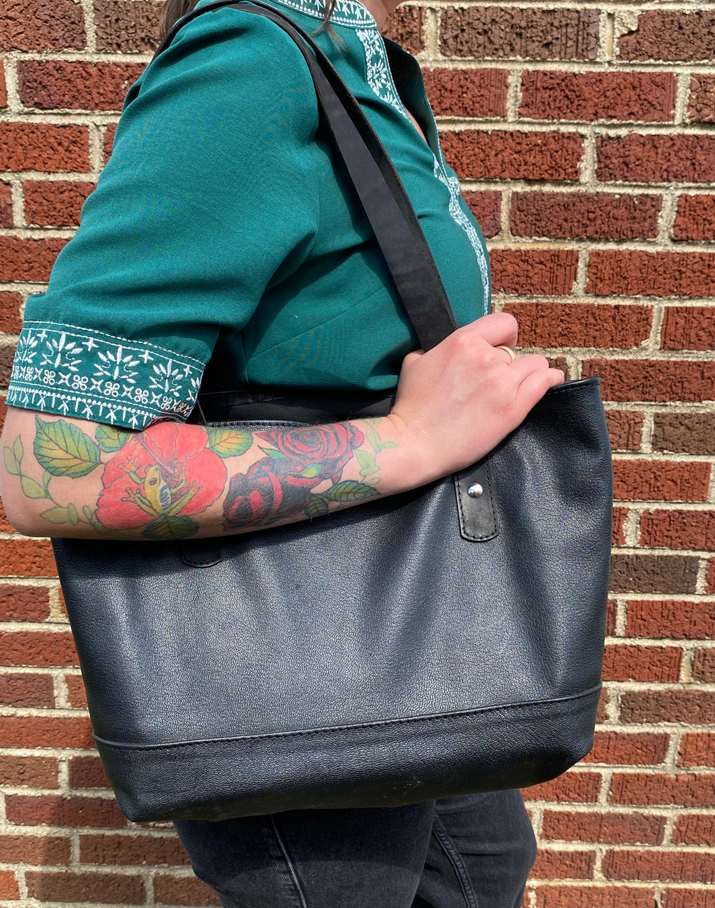 A woman wearing a green shirt and dark jeans carries a large black genuine Chevre goat leather tote bag with a shoulder strap, standing against a red brick wall.